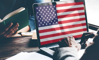 Männerhände vor Laptop, amerikanische Flagge auf dem ganzen Laptopbildschirm. Im Hintergrund liest eine Frau ein Buch.