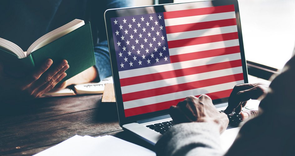 Männerhände vor Laptop, amerikanische Flagge auf dem ganzen Laptopbildschirm. Im Hintergrund liest eine Frau ein Buch.