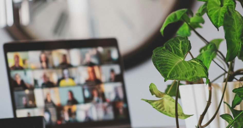 Laptop mit diversen Personen in einem Videoanruf rechts davon eine Pflanze dahinter eine grosse Uhr an der Wand